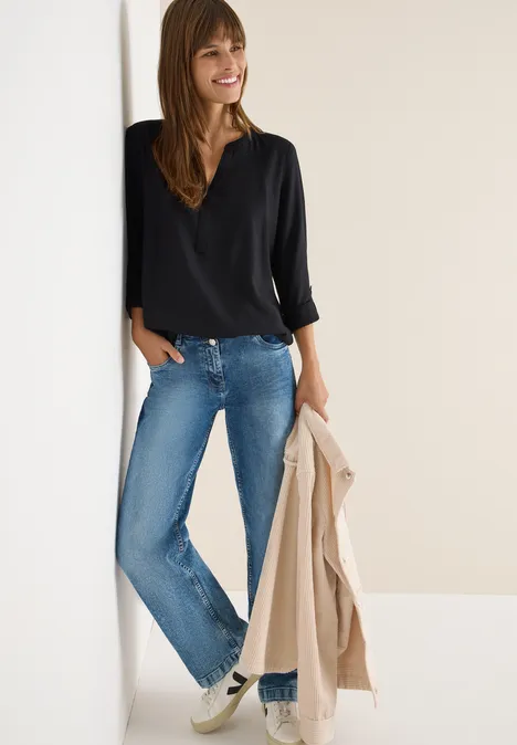 Frau mit schwarzem Oberteil, Jeans und Sneakers, hält eine Jacke, lehnt an der Wand und lächelt.